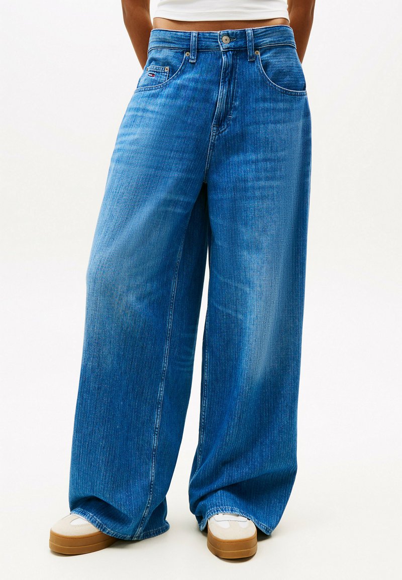 Weite blaue Denim-Jeans mit einem verwaschenen Look, Taschen vorne und einem Knopfverschluss. Kombiniert mit hellen Sneakers.