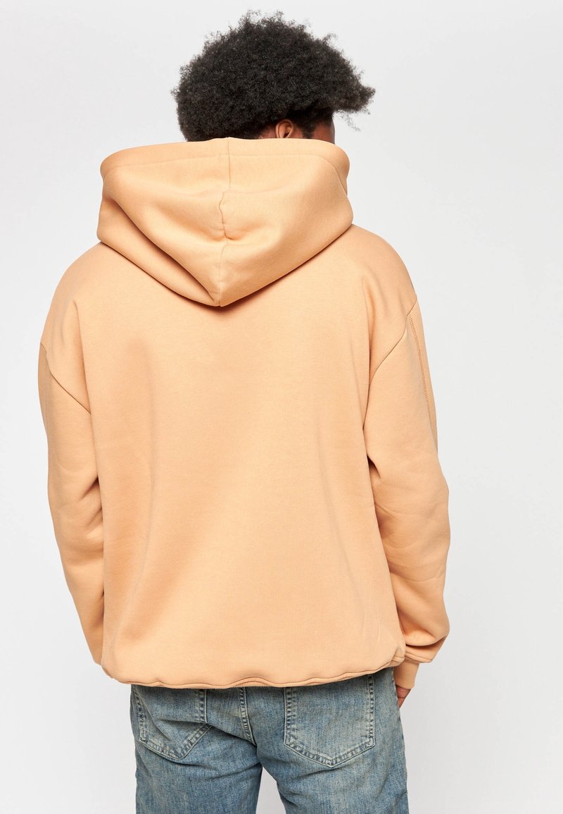 Personne aux cheveux bouclés portant un sweat à capuche pêche et un jean bleu, vue de dos sur un fond clair uni.