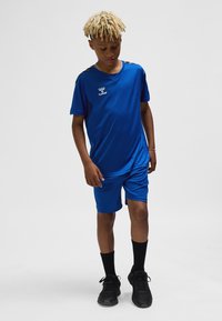 Blå sportoutfit i lätt material, bestående av en V-ringad tröja och shorts, med svarta detaljer på axlarna. Bärs med svarta strumpor och skor.