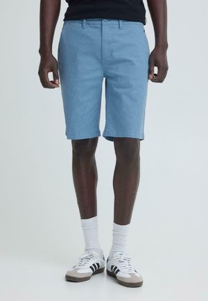 Mies, joka pukeutunut vaaleansinisiin shortseihin, valkoisiin sukkiin ja valkoisiin lenkkareihin, joissa mustat raidat, seisoo yksiväristä taustaa vasten.