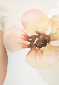 Κοντινή λήψη ενός λευκού πουκάμισου με μεγάλο, απαλό λουλούδι σε στυλ ακουαρέλας σε αποχρώσεις του ροζ, κίτρινου και καφέ.