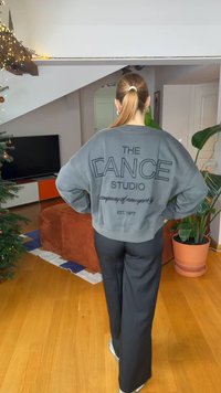 Γκρι βαμβακερό φούτερ με την επιγραφή «THE DANCE STUDIO» σε μαύρο χρώμα στην πλάτη· συνδυασμένο με μαύρο παντελόνι φαρδύ και λευκά αθλητικά παπούτσια.