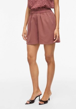 Vrouw draagt rosakleurige shorts met hoge taille, gecombineerd met een mouwloze top met structuur en zwarte open-toe muiltjes met hak, staand tegen een witte achtergrond.