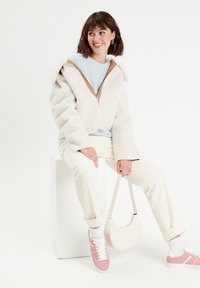 Chaqueta blanca de felpa con cremallera y grandes bolsillos, suéter azul claro, pantalones de pana color crema, zapatillas rosas y un bolso blanco.
