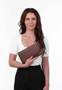 Hnědá kožená peněženka s elegantním designem a diagonálním klopou. Model je prezentován na jednobarevném pozadí, drží peněženku oběma rukama.