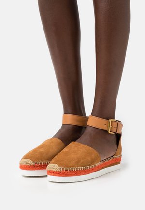 Chaussures style espadrille en daim beige avec détail en corde orange, semelle blanche et bride de cheville marron avec boucle dorée. Design à dos ouvert.