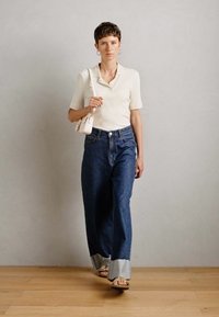 Ženska hodi oblečena v kremast kratkosleevni top, široke jeans hlače z raztrganimi robovi, bež sandale, uhane v obliki obroča ter nosi majhno kremasto torbico.