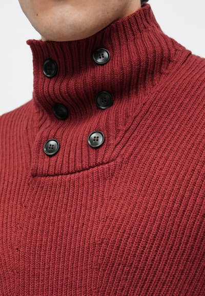 Knitted red sweater with a tall ribbed collar, featuring six black buttons and a textured pattern. Close-up shows stitching details.