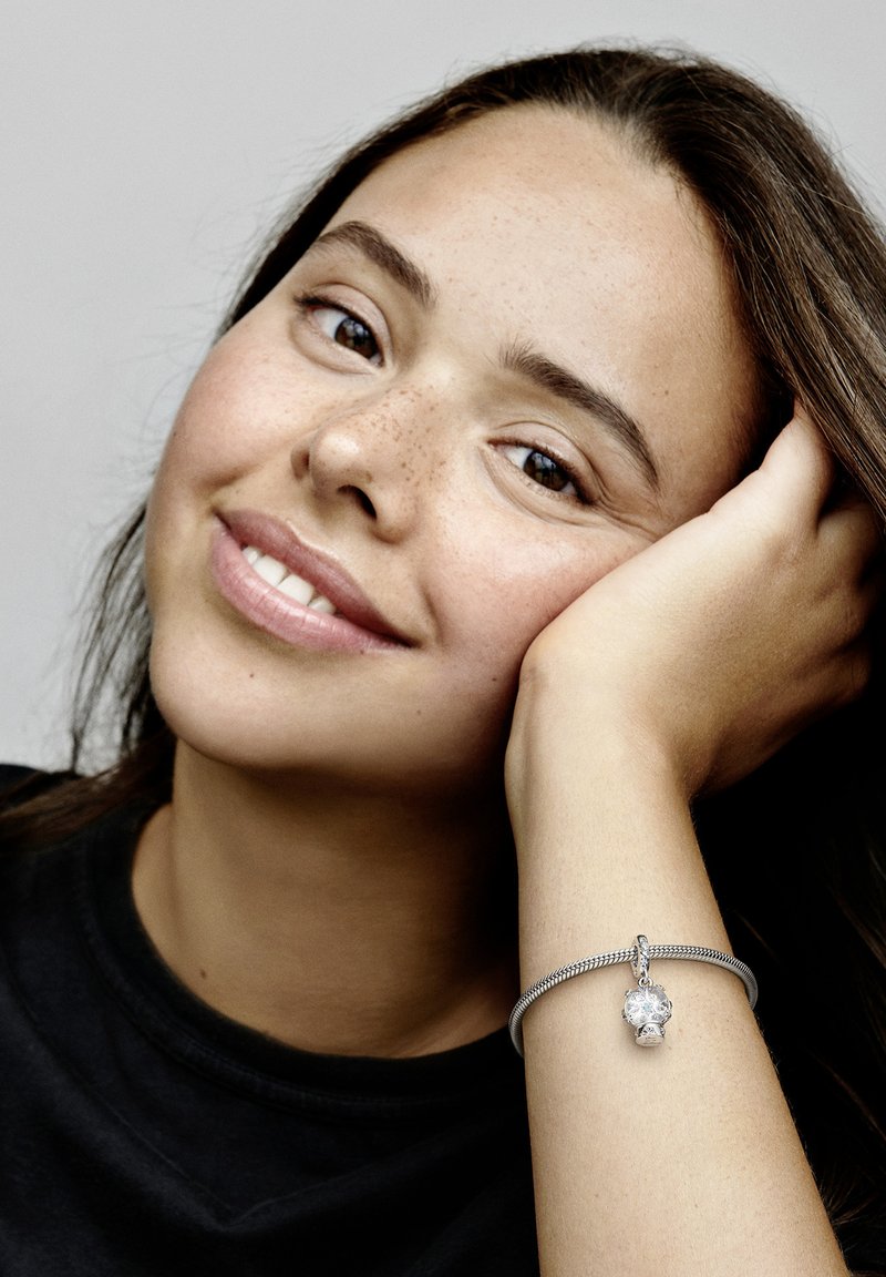 Srebrna zapestnica s teksturirano bandasom in prozornim kristalnim obeskom, nosi se na zapestju. Osvetljena polt in pegice, z črno majico.