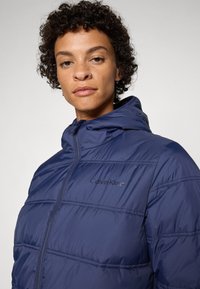 Veste matelassée bleu marine avec capuche, présentant des sections matelassées, une fermeture éclair à l'avant et un logo "Calvin Klein" brodé sur la poitrine. Texture lisse.