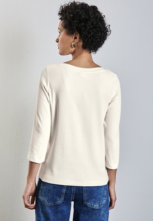 Haut beige clair à rayures, à manches longues avec un large décolleté et des manches trois-quarts, associé à un jean bleu foncé. Détails en tissu texturé visibles.