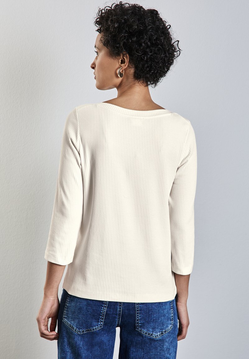 Helles beige, gestreiftes Langarmoberteil mit weitem Halsausschnitt und Dreiviertelärmeln, kombiniert mit dunkelblauen Jeans. Sichtbares Detail des strukturierten Stoffs.