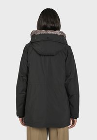 Svart parkasjacka med en huva fodrad med päls, med en avslappnad passform, synliga sömmar och en slät, matt textur. Stängning med knappar.