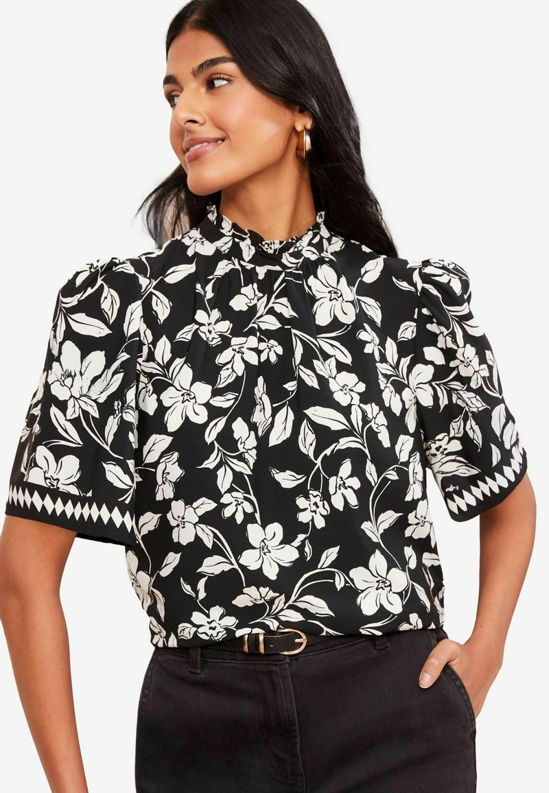 Schwarze Bluse mit weißem Blumenmuster, hohem Ausschnitt, Puffärmel und kontrastierenden Diamantakzenten an den Ärmelenden. Getragen mit schwarzen Hosen.