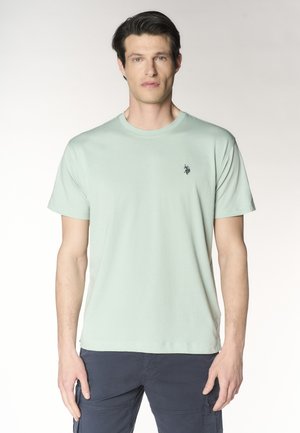 Uomo che indossa una maglietta verde chiaro a maniche corte con un piccolo logo nero sul petto e pantaloni cargo scuri, in piedi davanti a uno sfondo semplice.