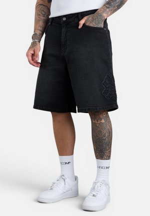 Mann trägt schwarze Jeansshorts mit besticktem Patch, weiße Socken und weiße Sneakers, zeigt tätowierte Arme und Beine.