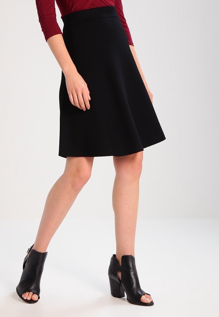 Femme portant une jupe noire trapèze, un haut bordeaux à manches longues et des bottines noires ouvertes à talons, debout, jambes croisées.