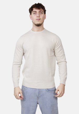 Giovane uomo in piedi, indossa un maglione beige a maniche lunghe e jeans azzurri chiari su uno sfondo bianco semplice.