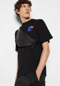Borsa a tracolla nera realizzata in materiale liscio, con una forma curvilinea, chiusura con zip e tracolla regolabile. Indossata sopra una t-shirt nera.