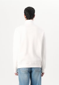 Vit sweatshirt med ribbad textur, hög krage och långa ärmar, åtföljd av ljusblå baggy jeans.