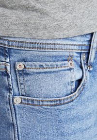 Prim-plan al unui buzunar de blugi denim de culoare albastru deschis, cu cusături vizibile și nituri metalice, purtați cu un top din țesătură gri.