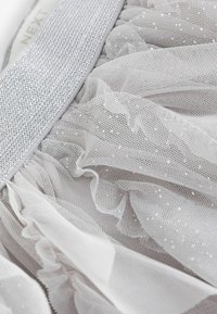 Tessuto di organza bianco con strati arricciati e accenti argentati. Presenta piccole paillettes riflettenti e una cintura argentata lucida.