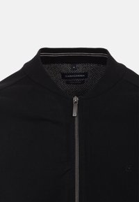 Musta vetoketjullinen takki, jossa on ribattu kaulus, sileä puuvillapinta ja kuvioitu sisävuori. Varustettu hopeanvärisellä vetoketjulla ja huomaamattomalla brändäyksellä.