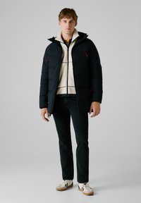 Chaqueta de plumón negra con acentos rojos, forro de felpa blanca, bolsillos con cremallera, llevada sobre una blusa amarilla, combinada con jeans oscuros y zapatos.