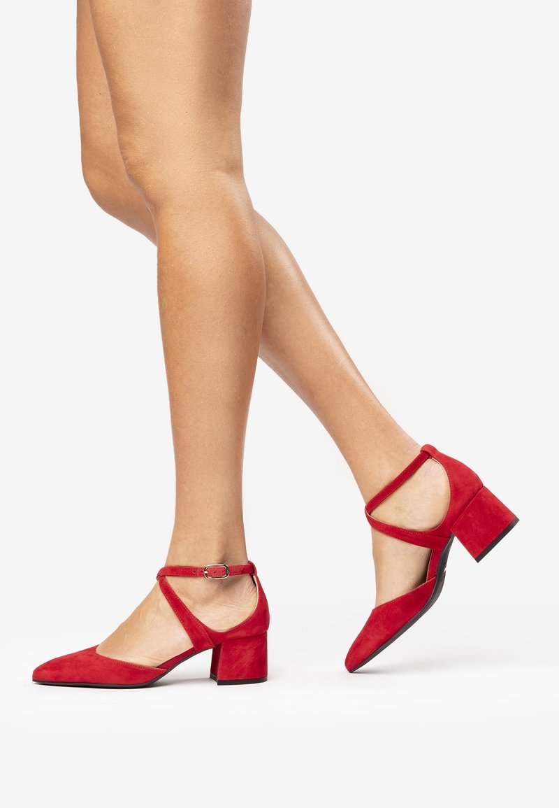 Jambes de femme portant des chaussures rouges en daim à talons blocs avec brides à la cheville sur fond blanc.