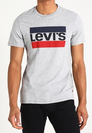 Mężczyzna w jasno szarej koszulce z krótkim rękawem z granatowym i czerwonym prostokątnym wzorem oraz wyraźnym czarnym napisem "Levi's" na przodzie.