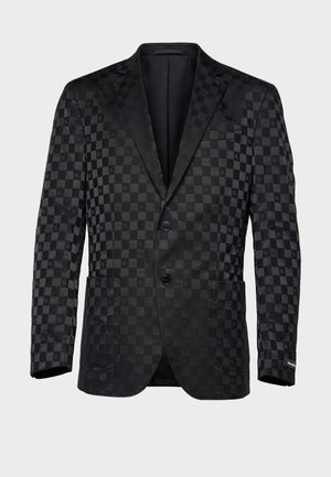 Un homme se tient devant un fond gris clair, vêtu d’une veste de costume sombre à carreaux, d’une chemise blanche, d’une cravate noire, d’un pantalon et de chaussures.