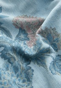 Tissu en denim bleu clair avec imprimé floral délavé dans des tons bleus et roses, montrant la texture et des plis doux.