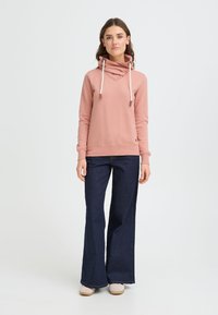 Sudadera con capucha rosa con cuello alto, cordones ajustables y dobladillo acanalado, combinada con vaqueros oscuros acampanados y zapatos beige sin cordones.