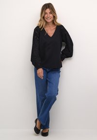 Schwarzes Oberteil mit V-Ausschnitt und Puffärmel im Lochmuster; kombiniert mit blauen gerade geschnittenen Jeans und schwarzen flachen Schuhen. Einfaches, lässiges Design.