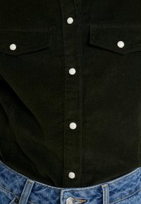 Mörkgrön kord skjorta med två bröstfickor, silverfärgade tryckknappar fram och en denimmidja synlig nedtill.