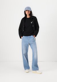 Svart jacka, svart t-shirt, ljusblå lösa jeans och vita sneakers. Ljusblå keps med logotyp på framsidan. Inomhus, enkel bakgrund.