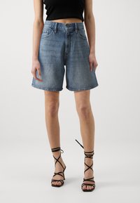 Högmidjade denimshorts i ljusblått med rak passform, försedd med framfickor och en fransad fåll. Matchas med svarta sandaler med remmar.