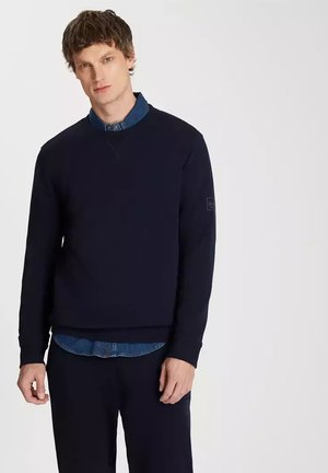 Navy blue sweatshirt in a soft fabric, featuring a crew neck and ribbed cuffs, layered over a denim shirt with a collar.