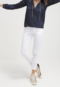 Mörkblå satinblus med en V-ringad hals och guldiga kedjeatt detaljer, ihop med vita denimjeans och guldiga sneakers med snördetaljer.