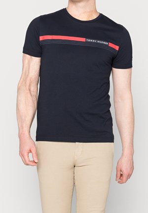Man wearing a fitted black Tommy Hilfiger t-shirt with red and black stripes across the chest, paired with beige pants.