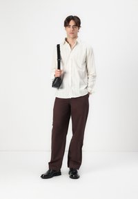 Hellgestreiftes Hemd, braune Hose, schwarze Schuhe und eine kleine schwarze Umhängetasche mit Kettenriemen. Das Outfit zeichnet sich durch eine lockere Passform aus.