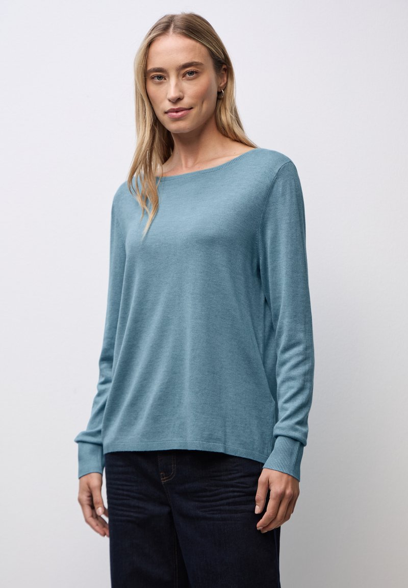Maglione a maglia azzurro chiaro con scollatura ampia, maniche lunghe e polsini a coste. Texture liscia, vestibilità rilassata, indossato con pantaloni in denim scuri.