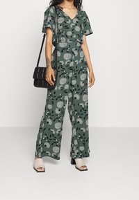 Blommig jumpsuit i grönt, med V-hals, korta ärmar, vida ben och ett svart blommönster. Matchad med en svart vadderad handväska.
