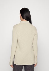 Beige långärmad topp i en texturerad tyg, med krage och sidoflikar. Inkluderar knappdetaljer på ärmarna, designad för en avslappnad passform.