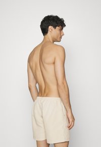 Smėlio spalvos maudymosi šortai su elastinga juosmens juosta, šoninėmis kišenėmis ir laisvu siluetu, demonstruojami ant nuogos odos vyriško modelio iš nugaros.