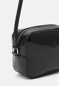 Borsa a tracolla nera in pelle verniciata con chiusura a zip e tracolla sottile e regolabile. Presenta una texture liscia e lucida e bordi arrotondati.