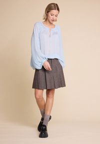 Kvinne iført en gjennomsiktig lyseblå bluse med puffede ermer, brun plisserte skjørt, grå strømper og svarte kraftige loafers på beige bakgrunn.