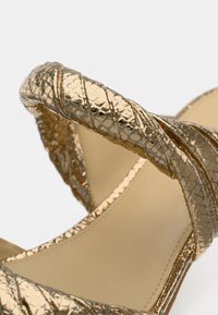 Πέδιλο με λουριά και ψηλό τακούνι σε μεταλλικό χρυσό, με ανάγλυφη υφή ερπετού, καμπύλο σχέδιο και λεία, εσωτερική επιφάνεια σε γυμνό χρώμα.
