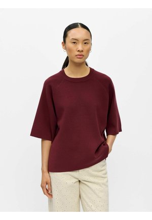 Femme aux cheveux foncés attachés, portant de petites boucles d'oreilles anneaux, un pull ample bordeaux à manches courtes et un pantalon beige clair à œillets, la main dans la poche.