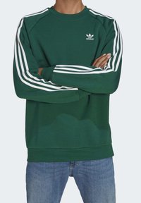 Grön sweatshirt i bomullsblandning med rund halsringning, prydd med vita tre ränder på ärmarna och en liten vit logotyp på bröstet.